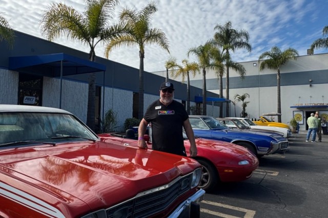 Man in black hat, sunglasses and black shirt, standing next to a classic red Pontiac car, with other classic cars parked behind him.